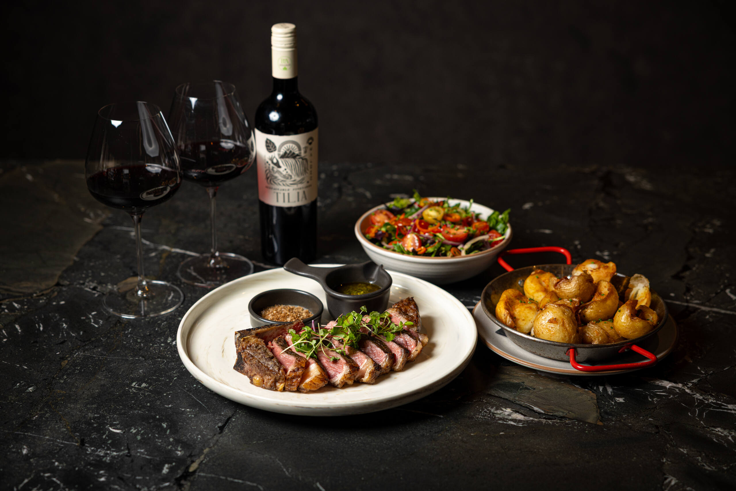 Image of a sirloin with potatoes, a side salad and a bottle of Malbec wine and two glasses at Calida.